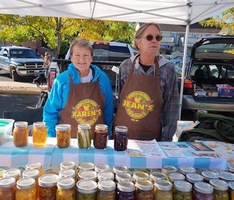 Karen Biesemeier and Jean Shanor in their aprons behind a market table lined with jars of pickles, jams and salsas